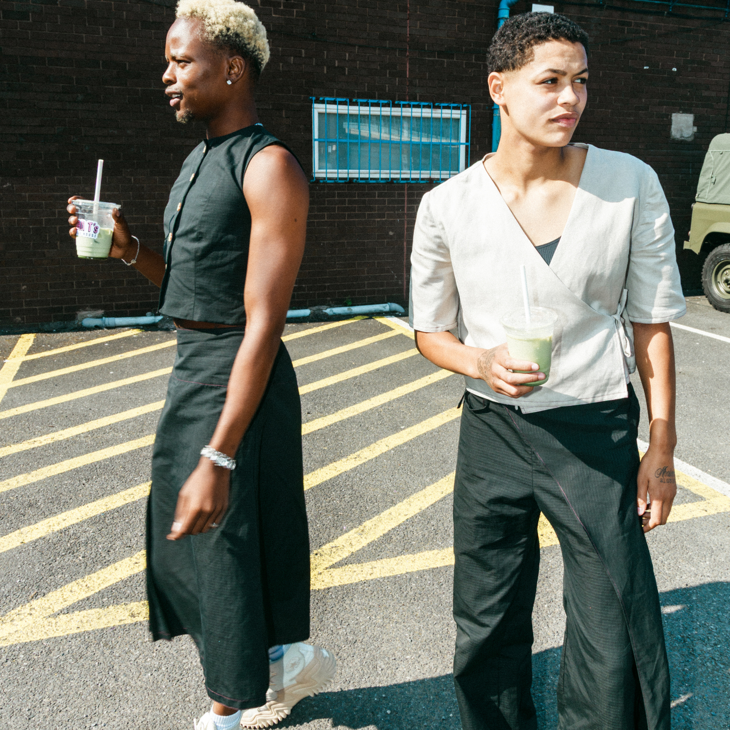 Two people walking outdoors, holding drinks, with a brick wall and parking lot in the background.