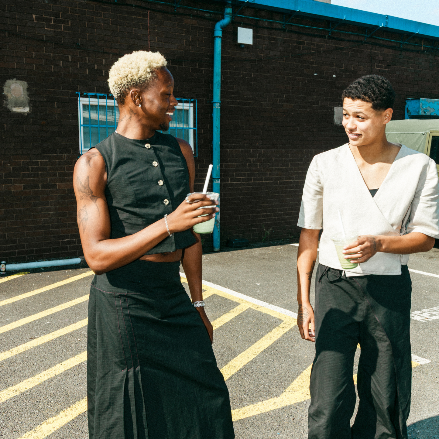 Two people standing in a parking lot, one holding a drink, against a brick wall.