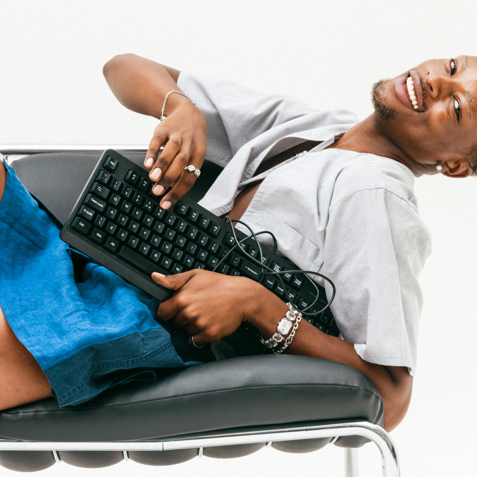 Person using a computer keyboard on a chair with a plain background wearing a tailored grey, upcycled shirt with a structured silhouette and a bow.