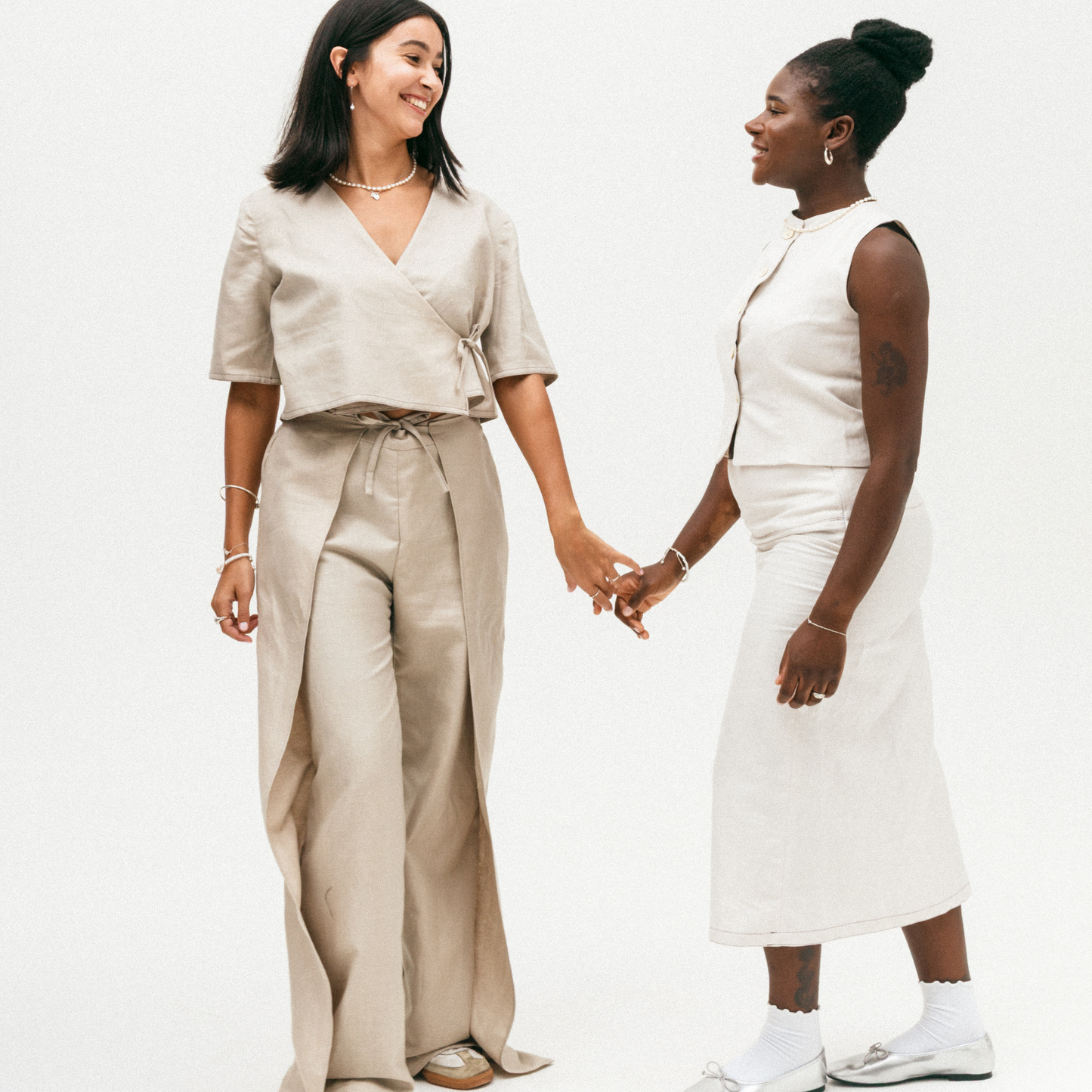 Two women holding hands, one in a beige outfit and the other in a white dress, on a plain background.