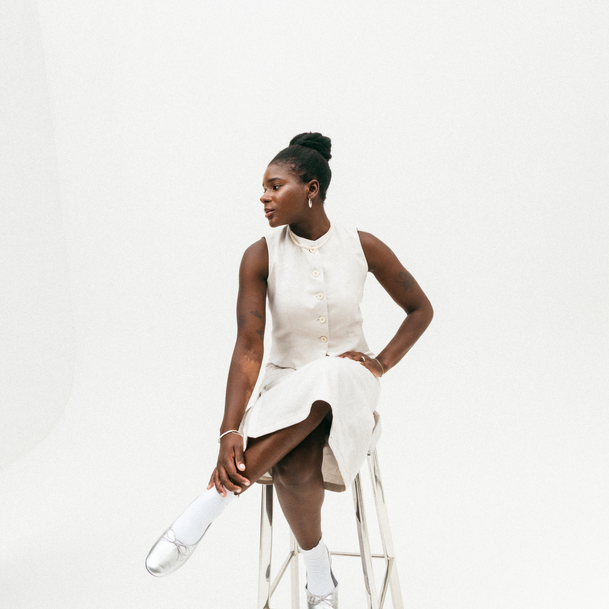 Woman in a white top and skirt sitting on a white stool against a white background