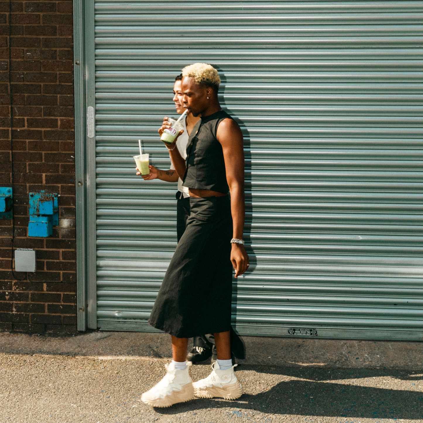 Person standing against a grey shutter holding a drink, wearing a black sleeveless top and skirt with white sneakers.