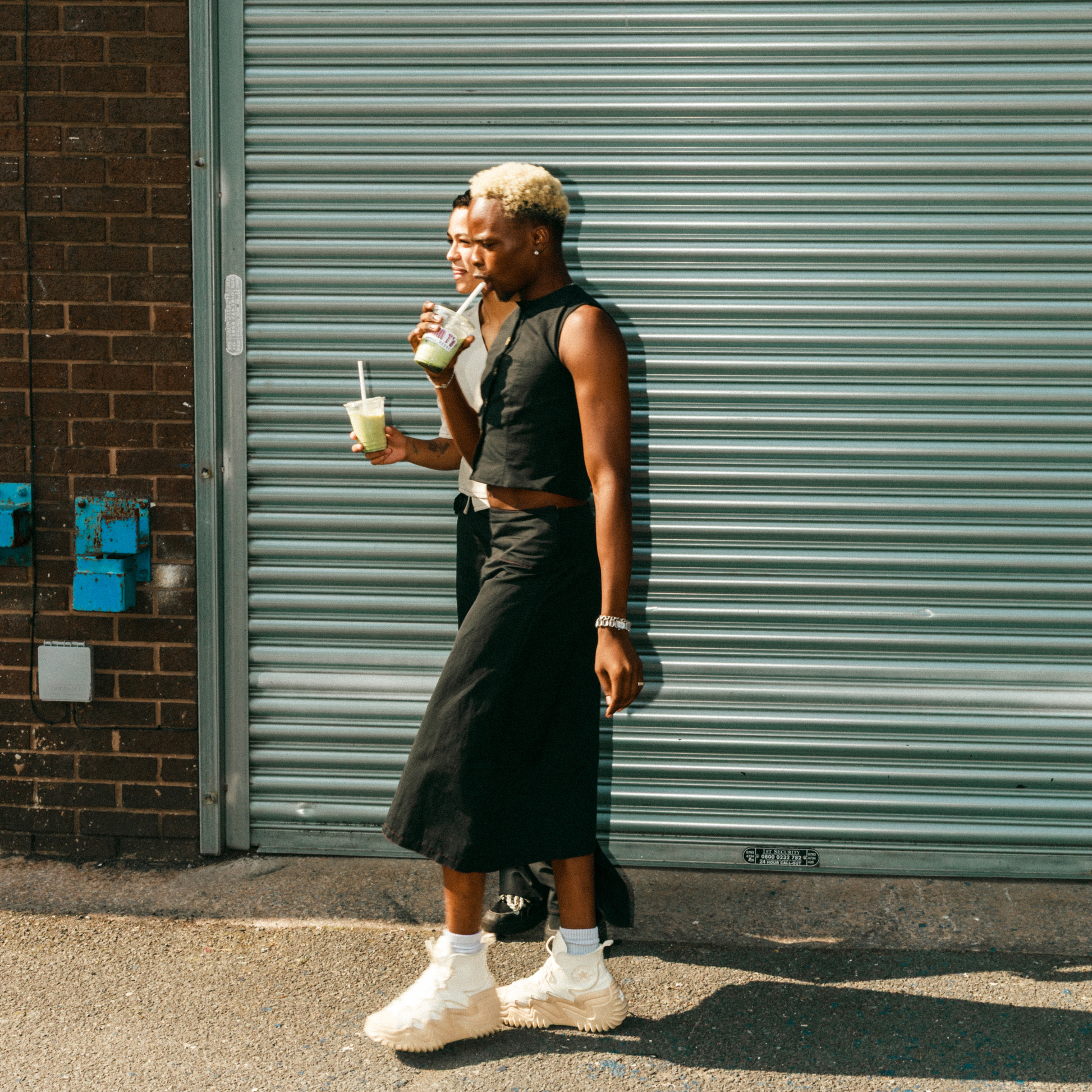 Person standing against a grey shutter holding a drink, wearing a black sleeveless top and skirt with white sneakers.