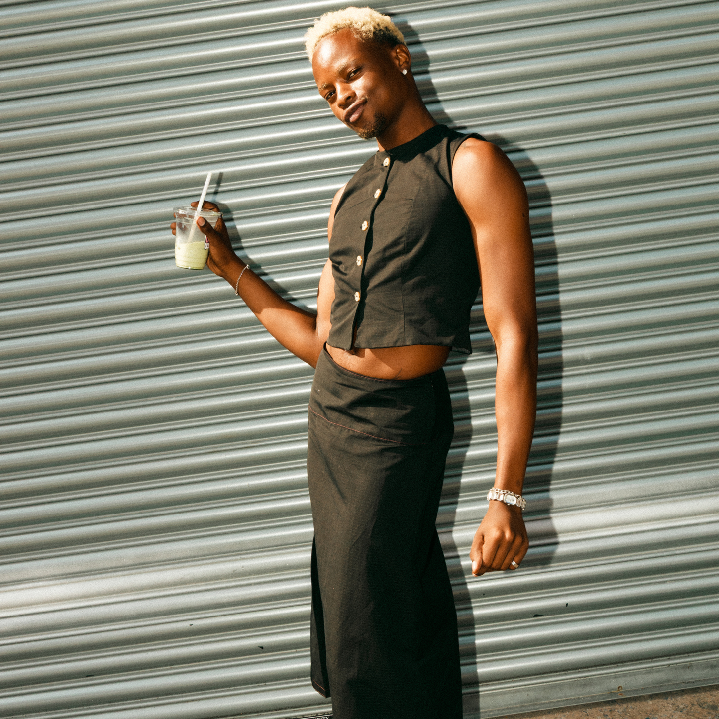 Person wearing a black sleeveless top and skirt holding a drink against a corrugated metal wall.