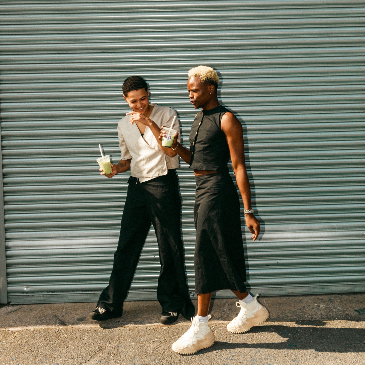 Two people standing next to a green metal shutter, holding drinks.