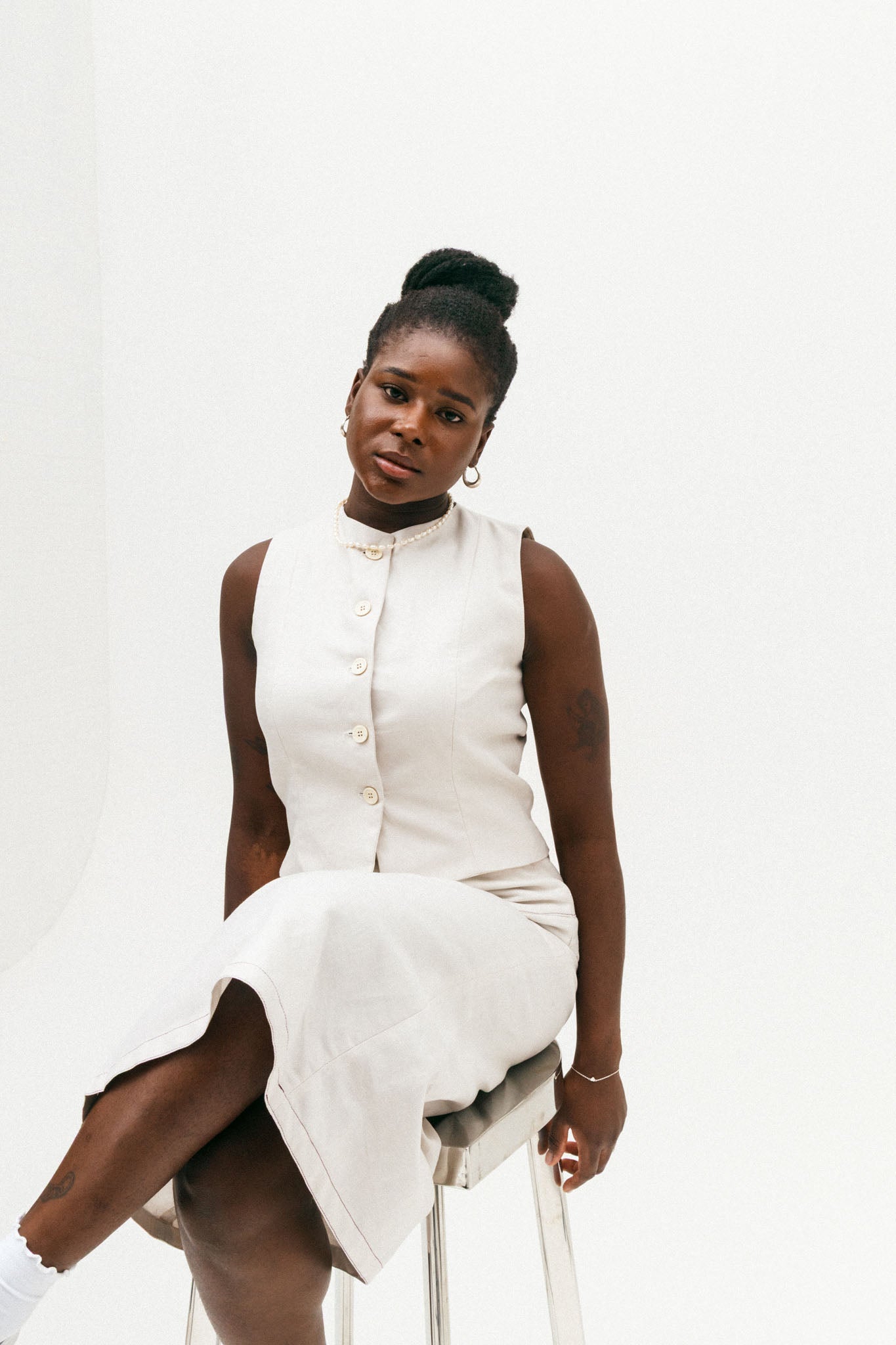 Woman wearing a white sleeveless dress sitting on a white stool against a white background